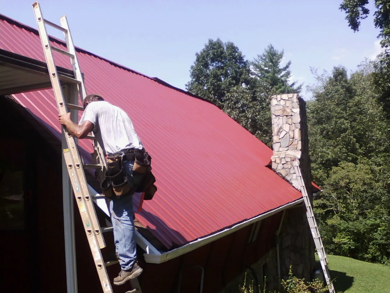 Roofer on a ladder beside a red metal roof and stone chimney — Soffit & Fascia Repair by Roofing Brothers in Twin Mountain